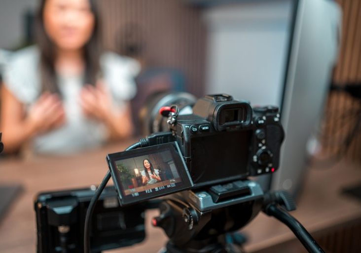 This striking photo features an Asian female content creator in a professional video recording setup. With a desktop PC and microphone, she confidently addresses the camera. The scene is elevated by meticulous lighting equipment placement.